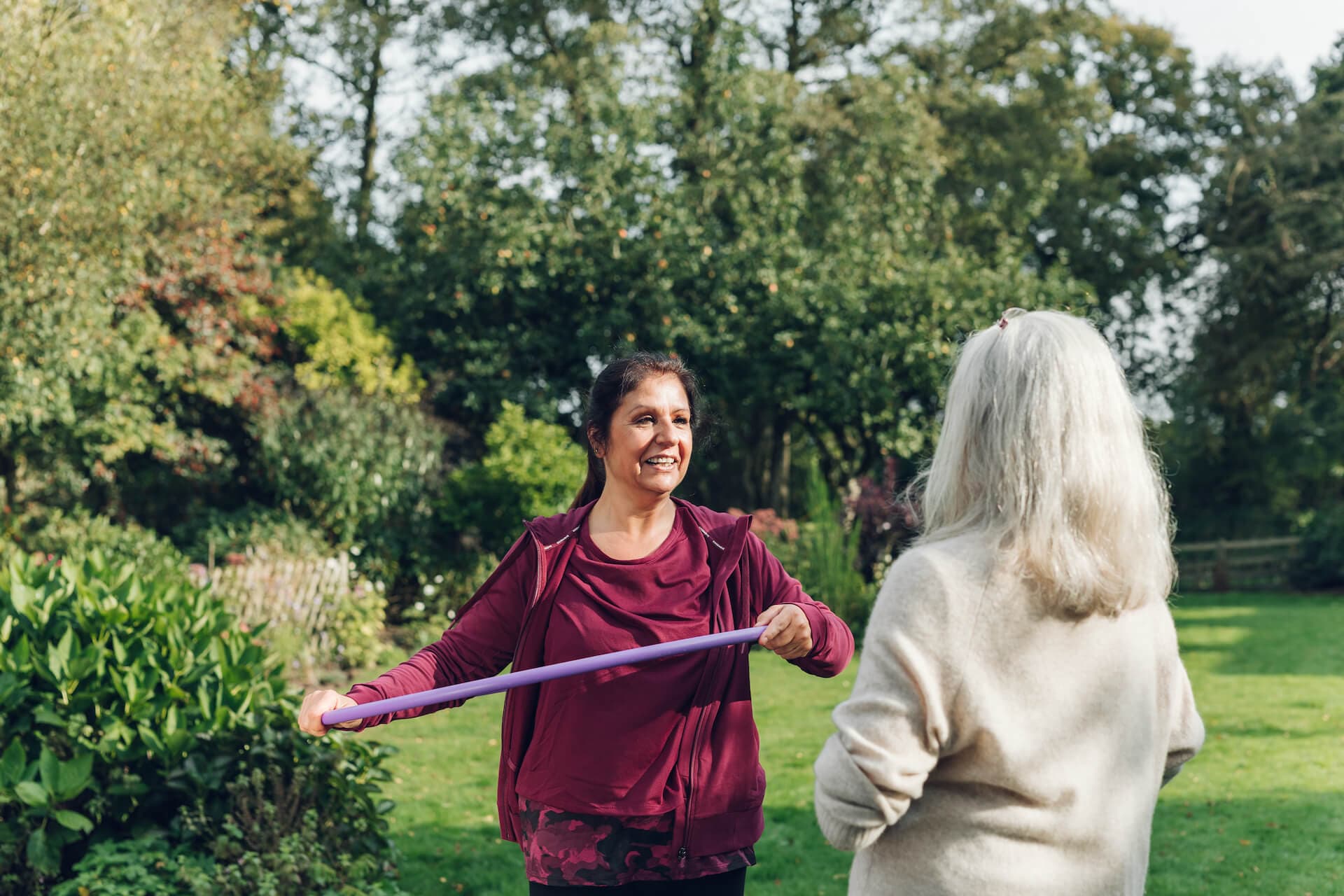 Two women in a garden, one holding a purple exercise band and smiling at the other, who has long white hair, trees in background. - Home Instead