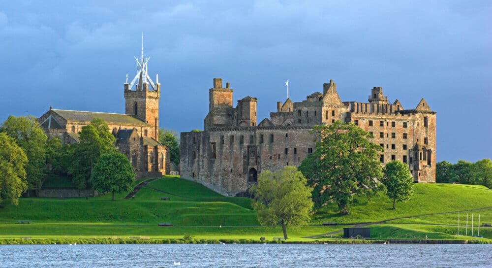 Historic stone castle with towers and green lawns, situated by a calm body of water under a cloudy sky. - Home Instead
