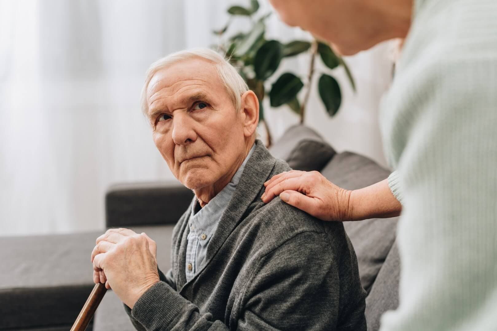 Elderly man sitting on a couch with a cane, looking up as another person gently places a hand on his shoulder. - Home Instead