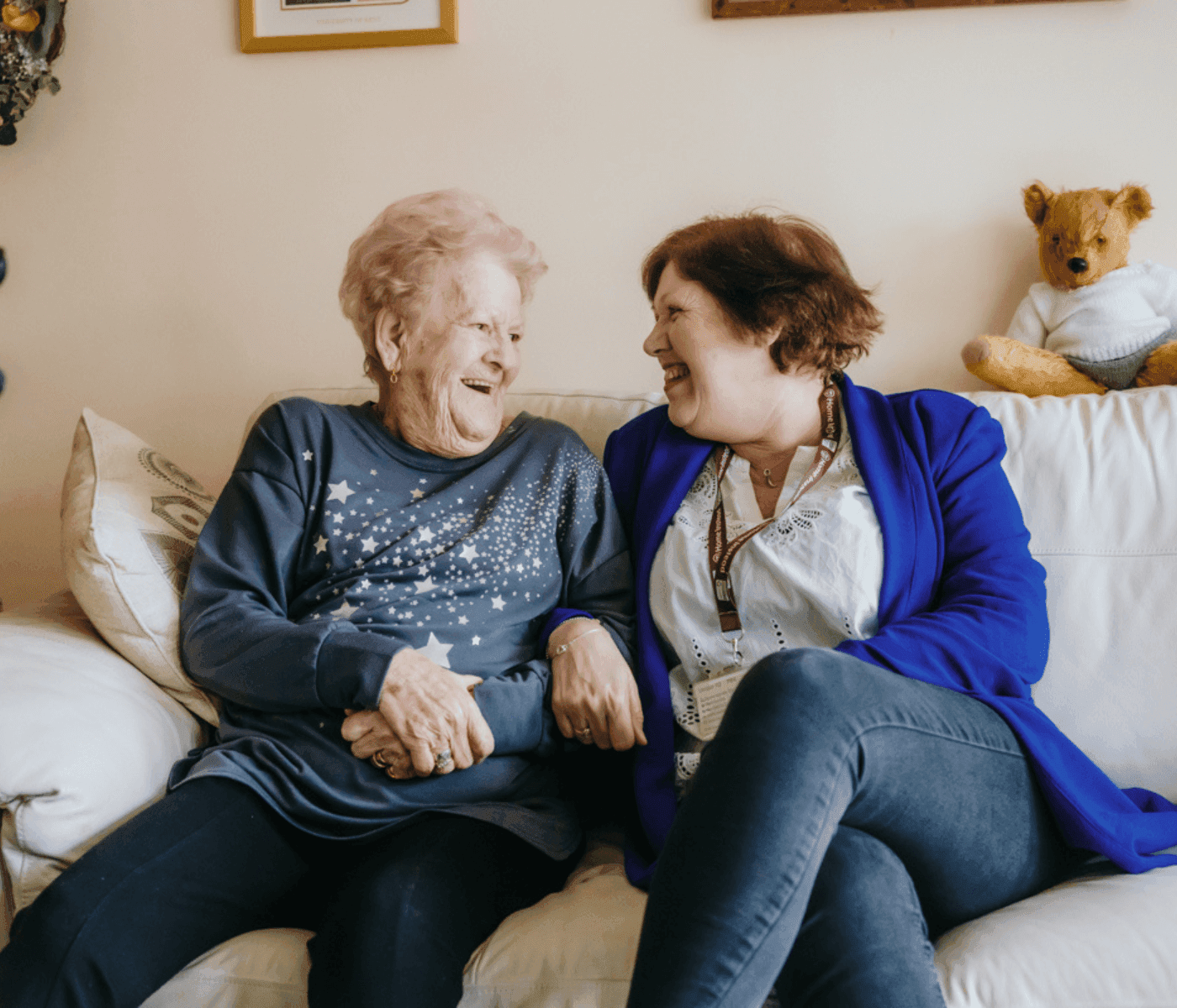 Two women sitting on a couch, smiling and holding hands, with a teddy bear in the background. - Home Instead