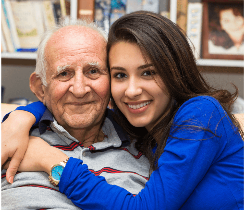A young woman in a blue top hugs an elderly man, smiling warmly in a cozy room with books and photos in the background. - Home Instead