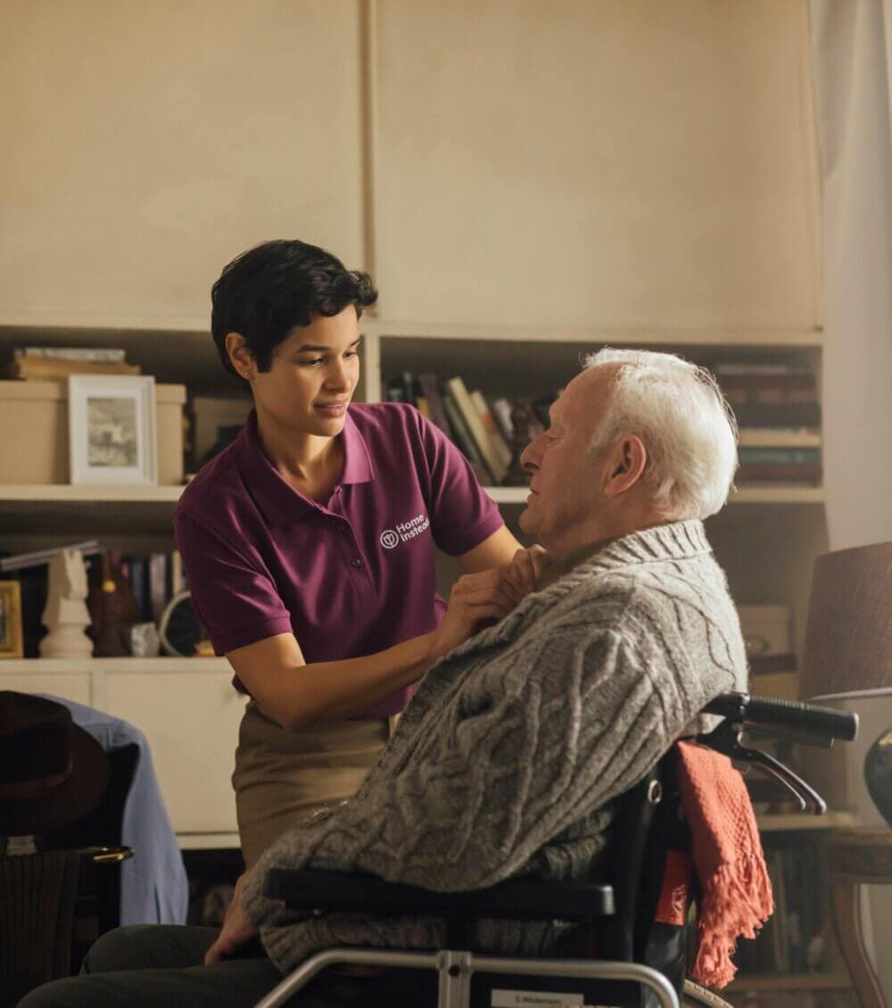 An elderly man sitting and smiling, being assisted by a caregiver who is also smiling. - Home Instead