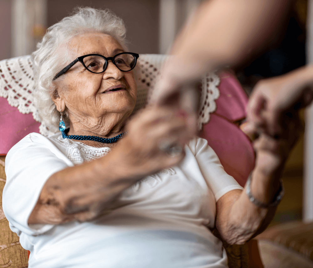Elderly woman with glasses sitting, smiling, and holding hands with someone. - Home Instead