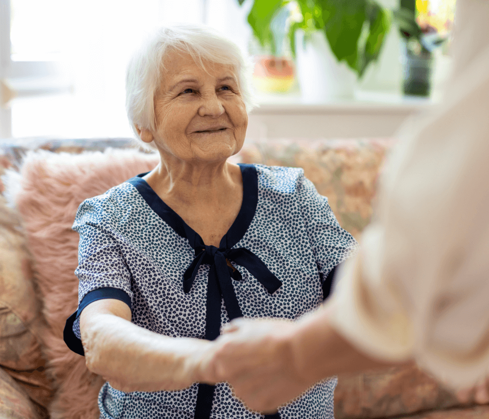 A smiling elderly woman with white hair shakes hands with another person while sitting on a couch. - Home Instead