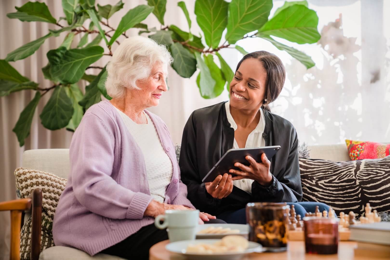 Elderly woman and younger woman smiling while looking at a tablet together in a cozy living room. - Home Instead