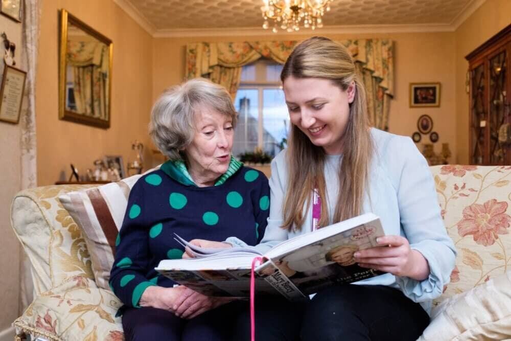 An elderly woman and a young woman sitting on a floral sofa, smiling while looking at a book together. - Home Instead