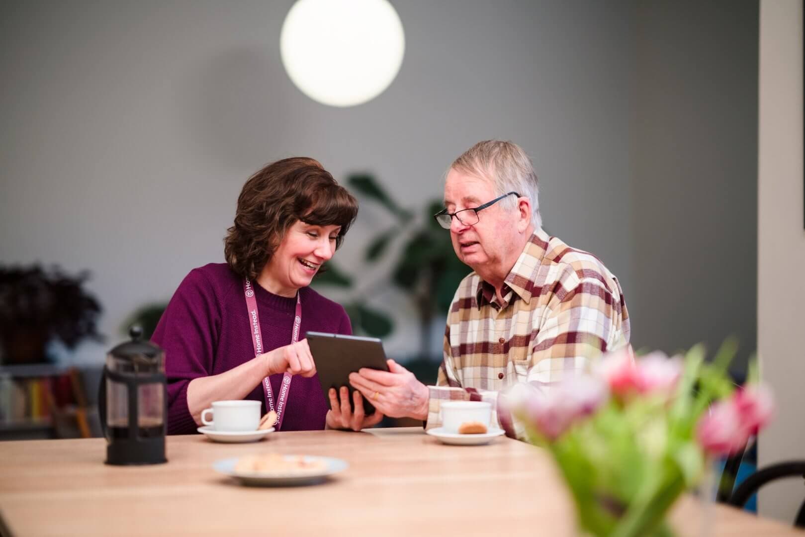 A woman and an older man sit at a table, smiling and looking at a tablet, with coffee and flowers in the foreground. - Home Instead