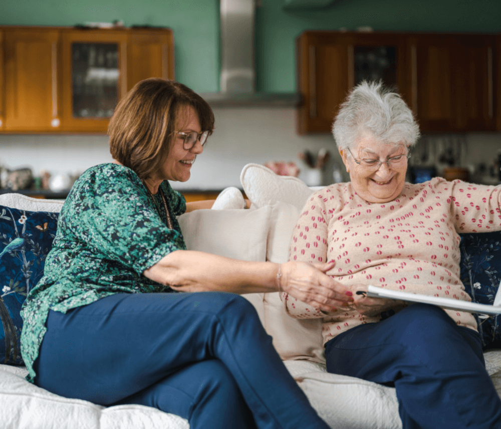 Two women sitting on a couch, smiling and looking at a photo album together in a cozy living room. - Home Instead