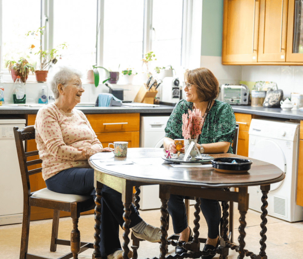 Two women smiling and chatting at a kitchen table with cups of tea and flowers on the table. - Home Instead