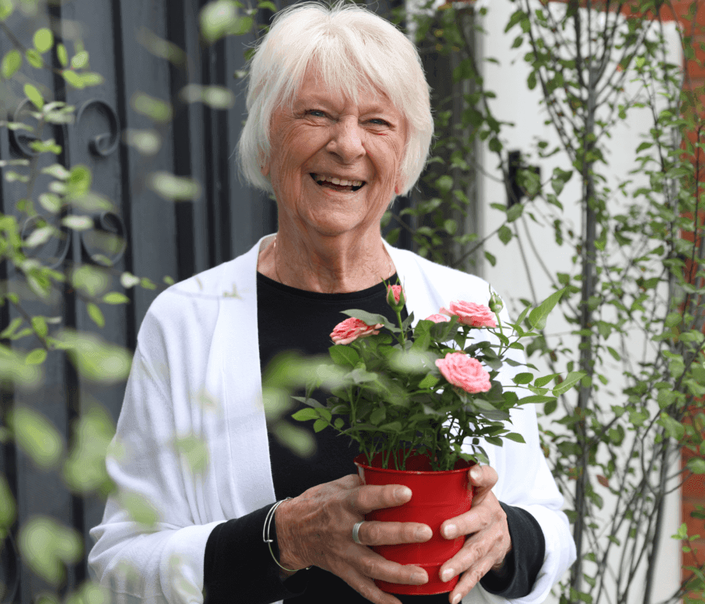 Elderly woman smiling and holding a red pot with pink flowers in a garden setting. - Home Instead