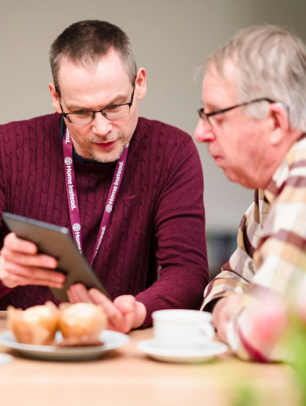 Two men sit at a table with coffee and muffins, one helping the other use a tablet. - Home Instead
