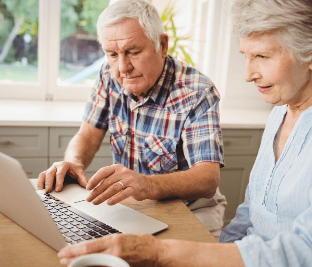 Elderly couple using a laptop together at a kitchen table, with a window in the background. - Home Instead