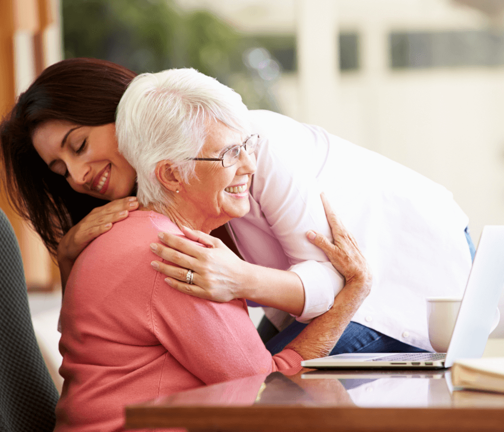 A younger woman hugs an older woman who is sitting and smiling in front of a laptop. - Home Instead
