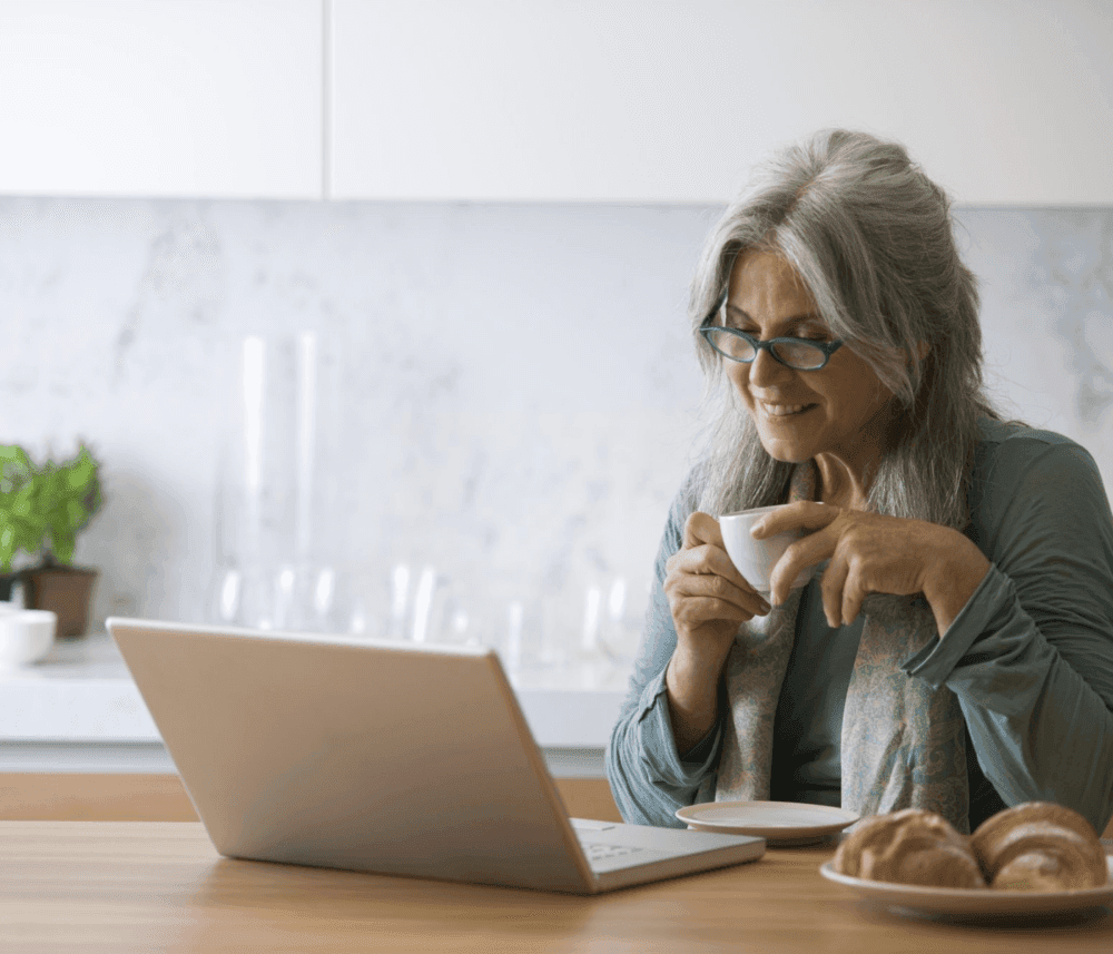 Older woman with gray hair and glasses, smiling, drinking coffee, looking at laptop, sitting at a table with pastries. - Home Instead