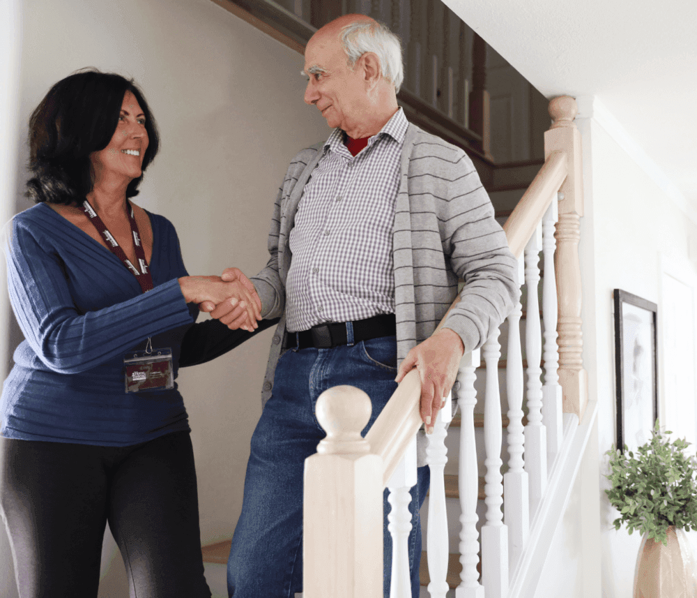 A woman shakes hands with an elderly man at the bottom of a staircase, smiling warmly at each other. - Home Instead