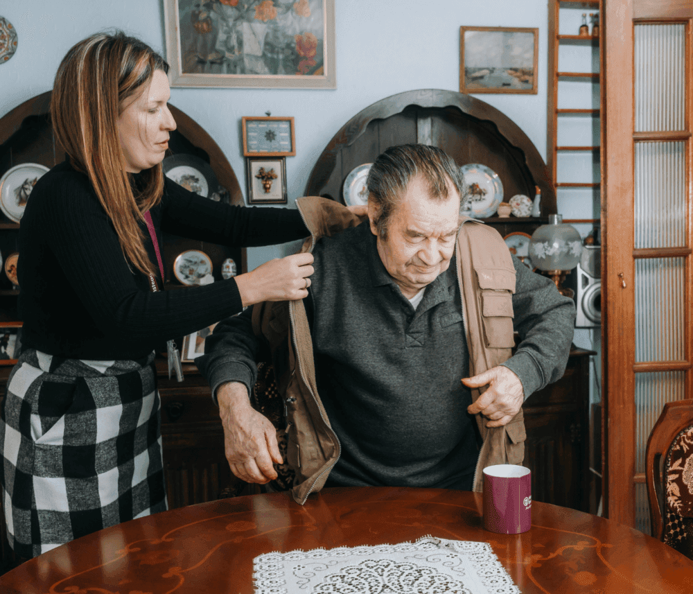 A woman helps an elderly man put on a jacket while he sits at a table with a mug. - Home Instead