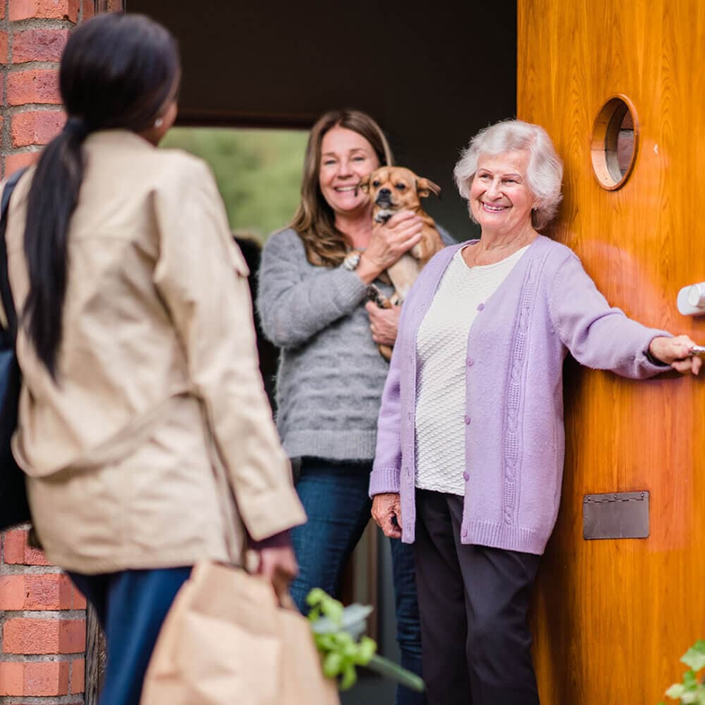 An elderly woman and a younger woman with a dog welcome a visitor at the front door of a home. - Home Instead