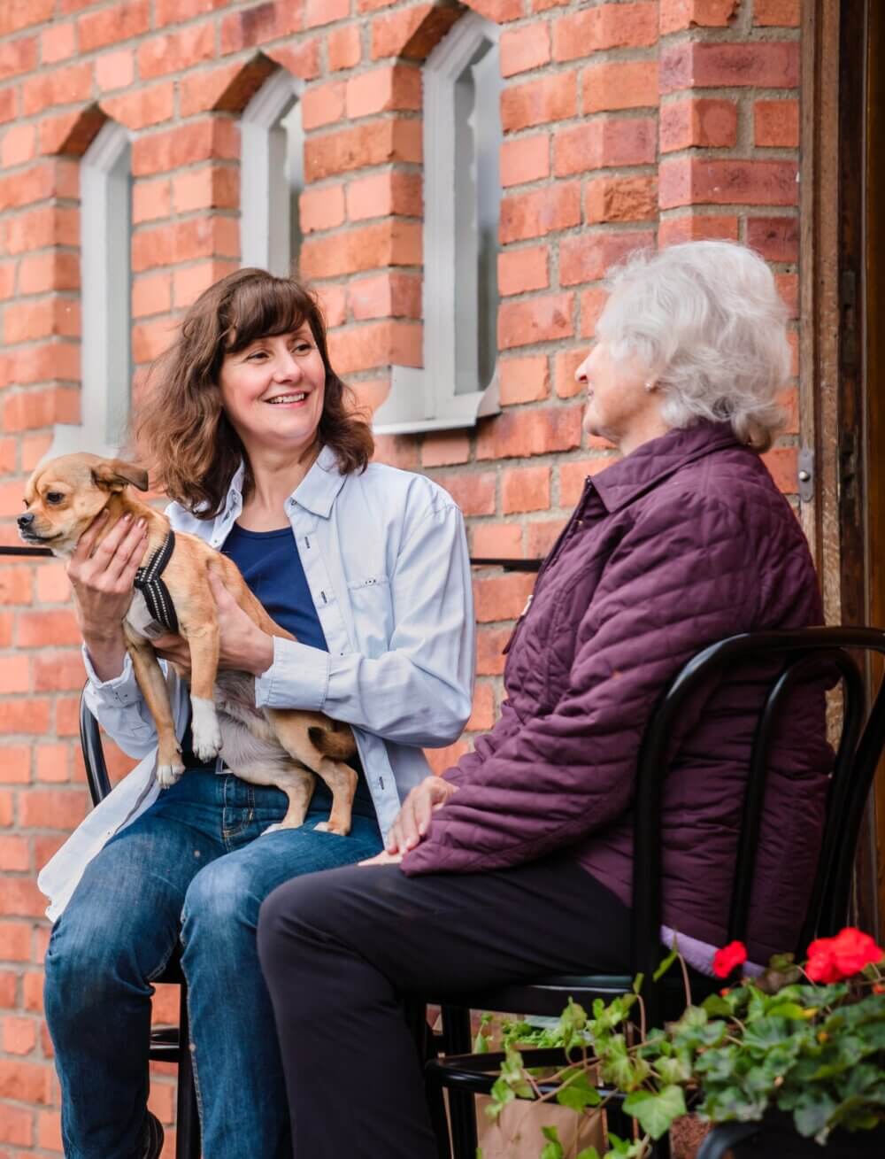 Two women, one holding a small dog, sit outside a brick building, smiling and chatting together. - Home Instead