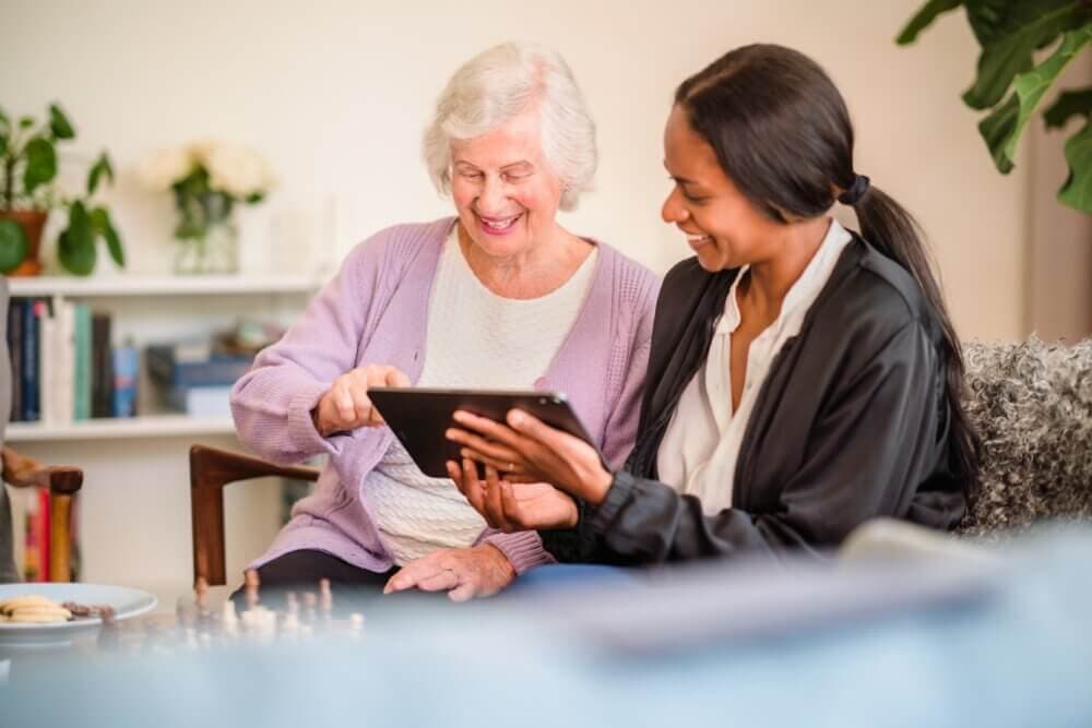 Elderly woman and young woman smiling while using a tablet together in a cozy, well-lit living room. - Home Instead