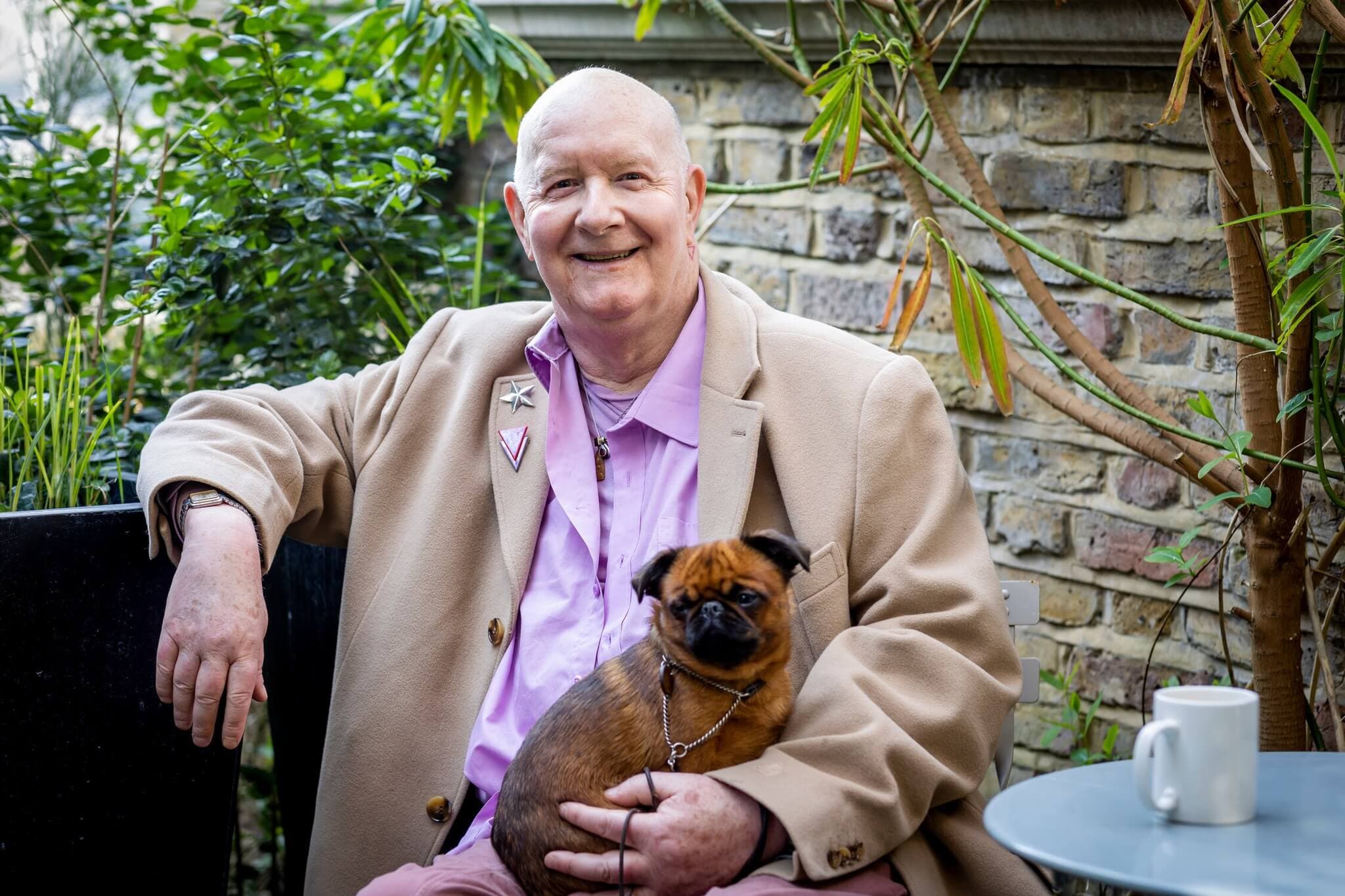 Man in a beige coat and pink shirt sits outdoors with a small dog on his lap, smiling; a coffee cup beside him. - Home Instead