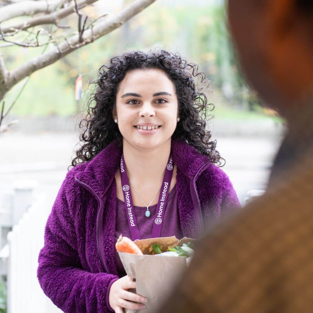 A Home Instead Care Assistant with curly hair in a purple jacket holding a grocery bag at a doorstep, smiling warmly. - Home Instead
