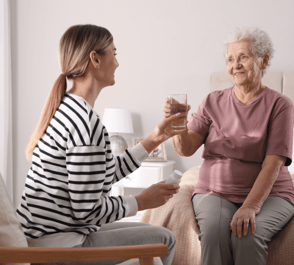 A young woman hands a glass of water to an elderly woman sitting on a bed in a cozy bedroom. - Home Instead