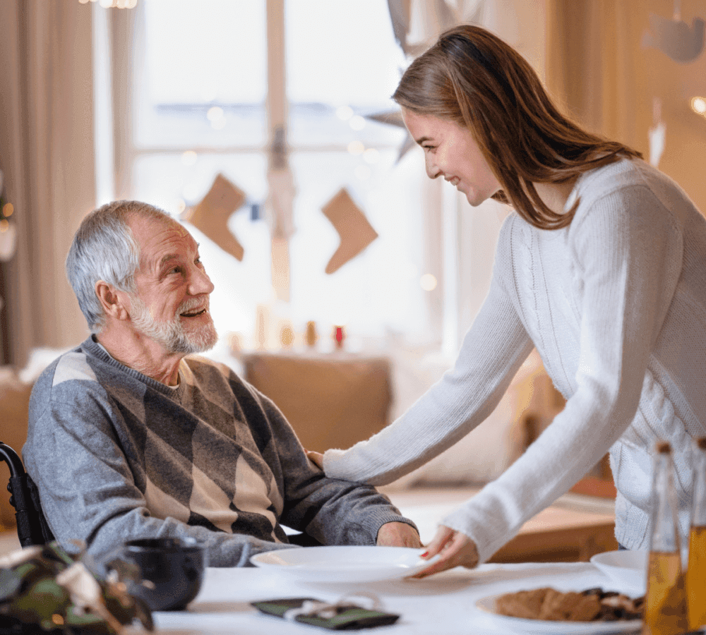 A young woman smiles while helping an elderly man in a wheelchair at a festive, warmly lit dining table. - Home Instead