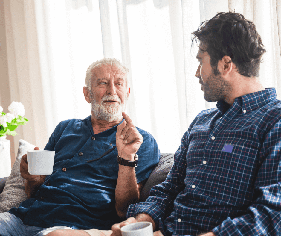 Older man and younger man sitting on a couch, having a conversation while holding white mugs, in a bright room. - Home Instead
