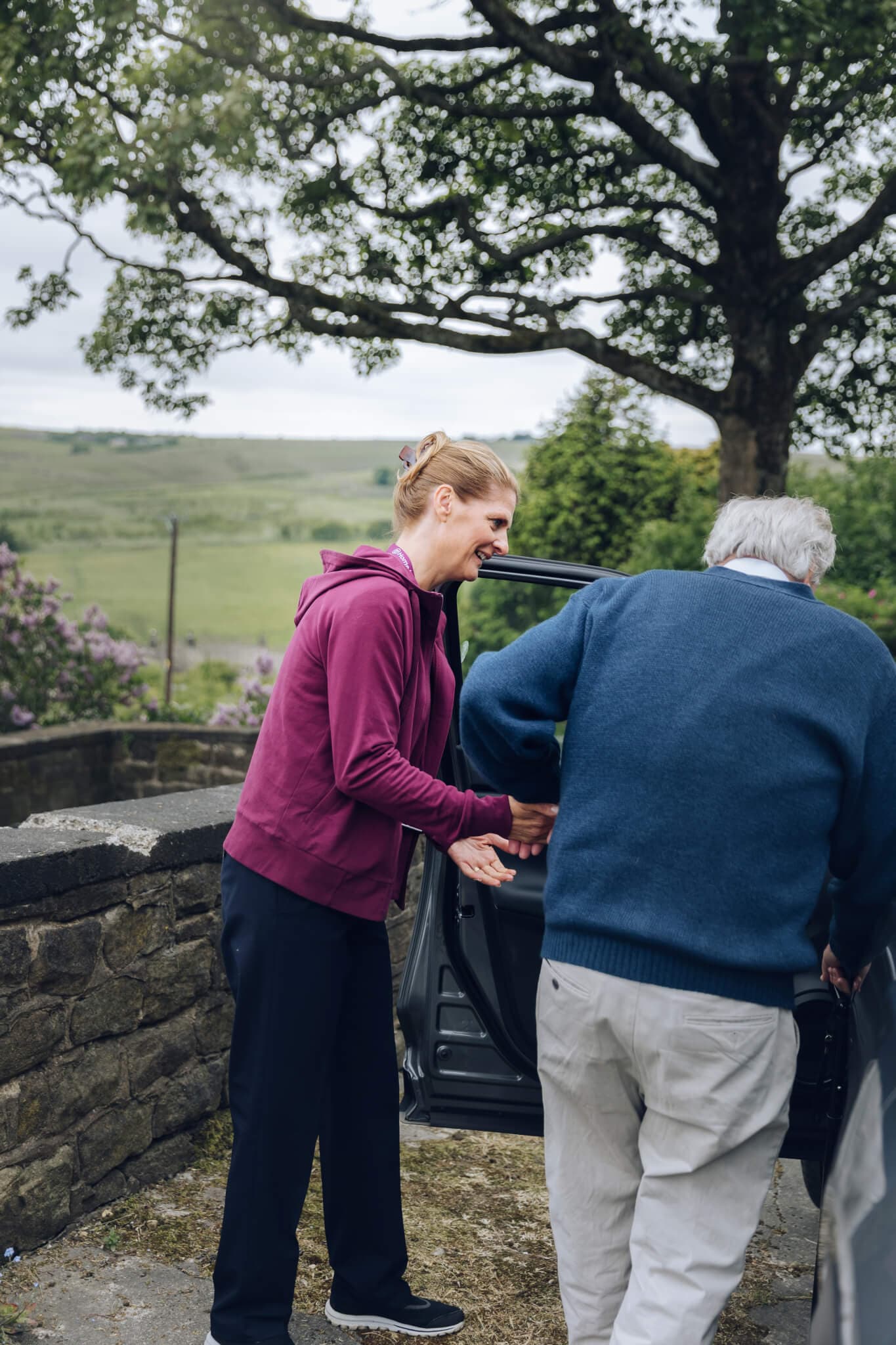 A woman helps an elderly man get out of a car in a scenic, rural area with trees and fields in the background. - Home Instead