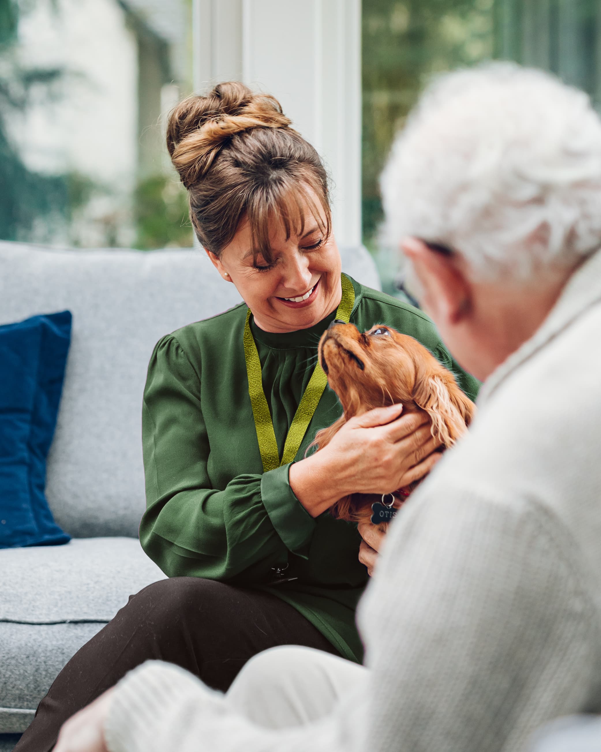 A woman and an elderly man sit on a couch, smiling as the woman holds a small dog. - Home Instead