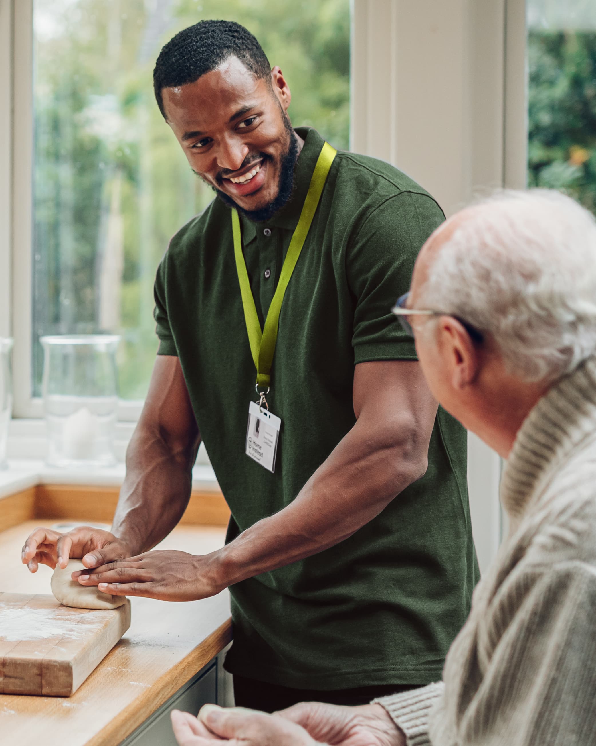 A smiling caregiver helps an elderly man knead dough in a bright kitchen. - Home Instead