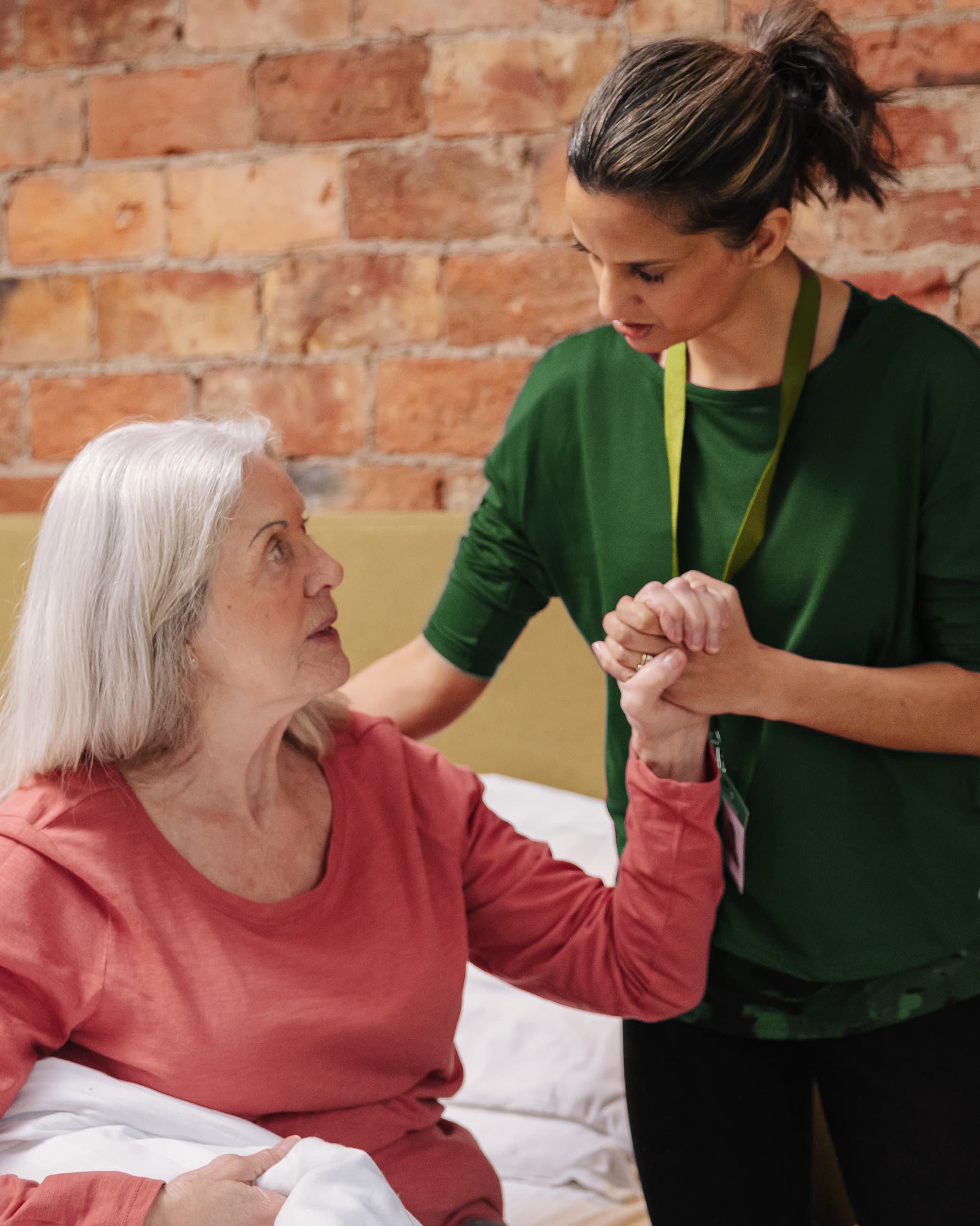 A caregiver in green supports an elderly woman in pink, holding her hand beside a bed. - Home Instead